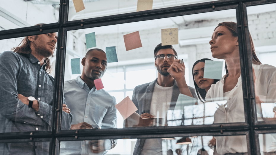 A group of young professionals stand in front of a large window where they have placed sticky notes. They are all looking with interest at them and appear to be making a business decision together. One woman has a pen and appears to be leading the meeting.