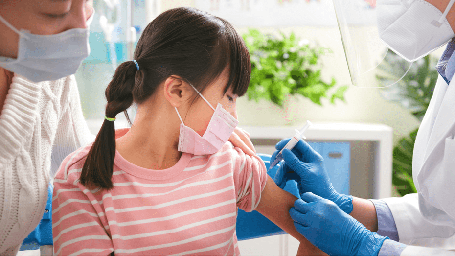 Doctor wearing gloves and mask administers a COVID-19 vaccination to a child patient as her mother observes.