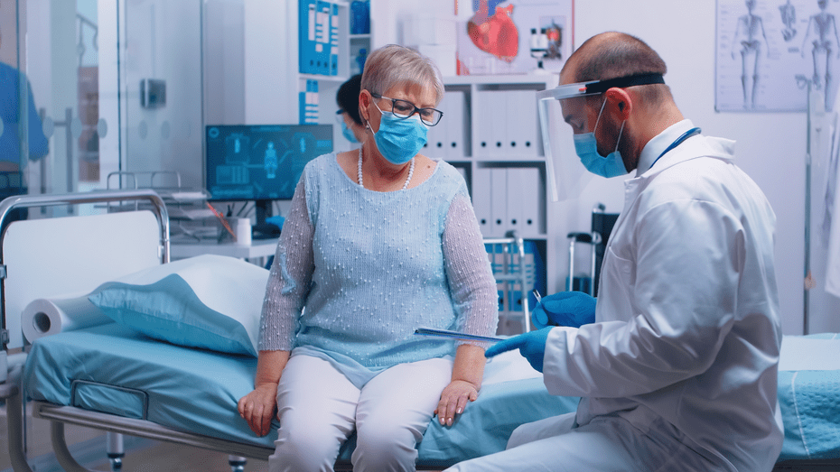 A gray-haired woman wearing a blue surgical mask sits on a hospital bed and consults with a provider wearing a white coat, blue mask, and face shield.