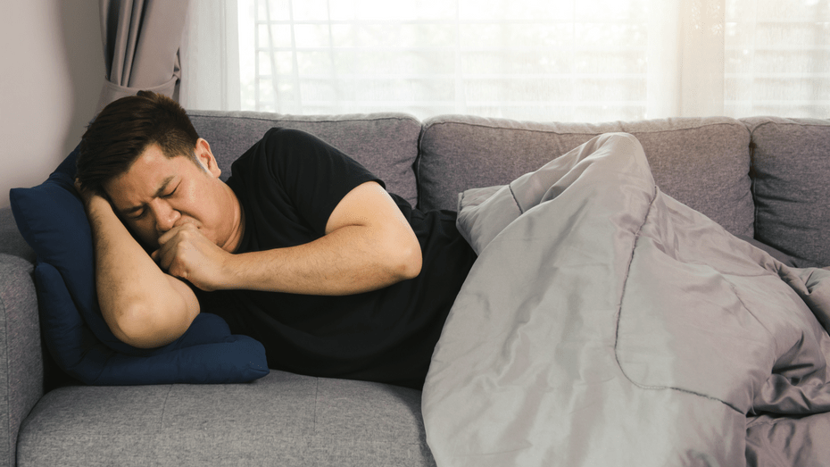 An Asian man lies on a gray couch under a gray quilt. He is resting his arm and head against a pillow and sitting up slightly to cough. 