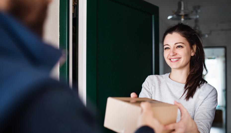 A white man in a blue zip-up hands a prescription delivery in a brown box to a smiling white woman with dark hair and a white shirt.