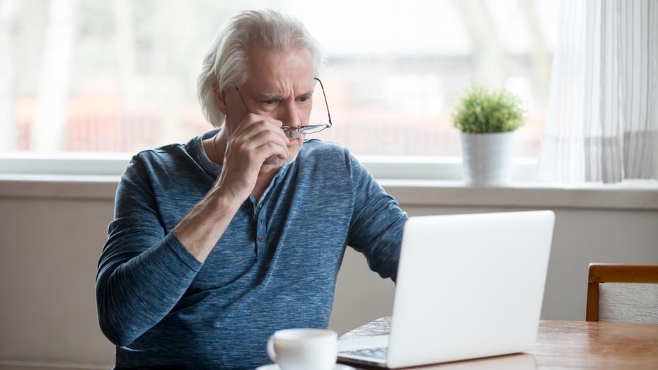 An older man in a blue shirt puts on his glasses as he reads something on a laptop. 