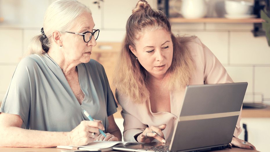 Two white women sit at a kitchen table, looking at a laptop. On the left, an older woman with white hair and glasses holds a pen over a notebook, ready to take notes. On the right, a middle-aged woman with brown hair leans towards the computer, typing with one hand. 