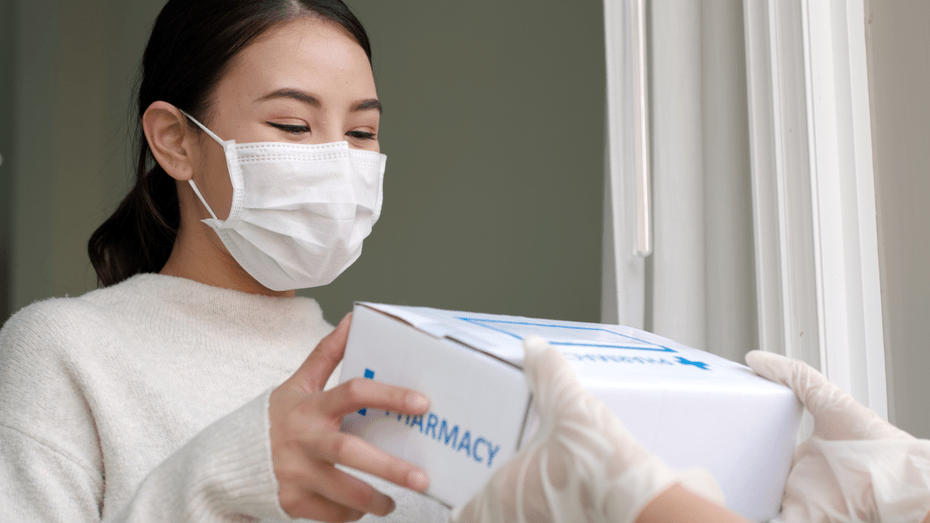 A woman on left of image with dark hair and white mask and shirt accepts a pharmacy shipment from a person wearing latex gloves who is outside of frame.